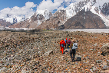 A group of hikers walks on a rocky trail with a base camp and a majestic snowcapped mountain range in the background on a sunny day