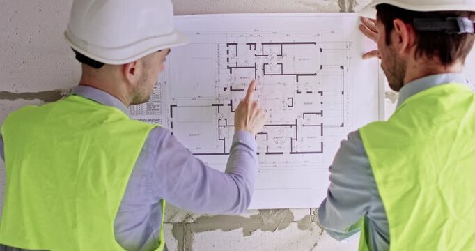 Specialists unfold paper sheet with floor plan of apartment put on wall. Male masters analyze stages of future work using large visual scheme standing by cinder blocks