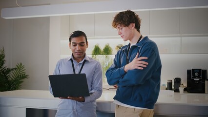 Two diverse people in modern startup office. thoughtful highschool student or intern gets mentorship while looking at laptop with colleague during project discussion