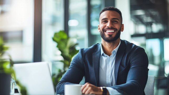 A smiling businessman sitting at a desk with a laptop and a cup of coffee in an office setting.