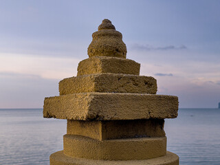 A weathered, tiered stone pillar with a rounded top stands against a calm sea and a soft twilight sky, showing its textured surface in fading light.