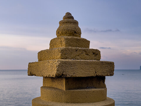 A weathered, tiered stone pillar with a rounded top stands against a calm sea and a soft twilight sky, showing its textured surface in fading light.