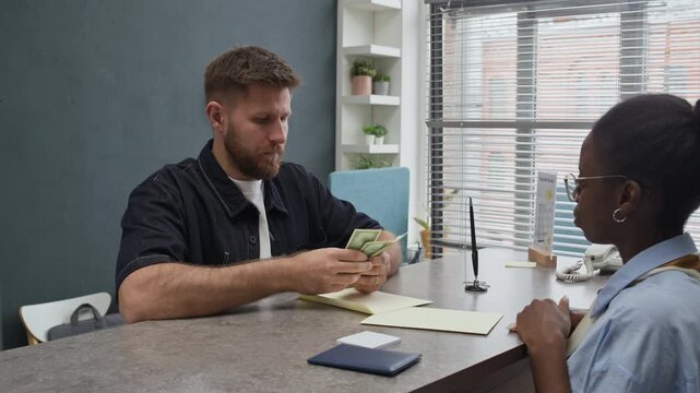 Handheld shot of female black teller handing banknotes in envelope to male Caucasian client counting in bank office