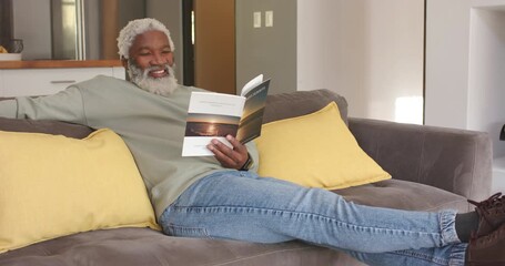 After opening paperback book Senior African American man reading and turning pages to relax on sofa