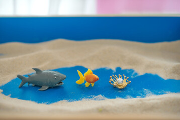 The concept of psychology. Sand
therapy. Shark figurines swimming after a goldfish, on the sand in a blue sandbox. Psychology. Therapy. PTSD. Confrontation. Aggression.