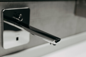 A modern chrome faucet in a luxury bathroom, with a sensor for touchless use. Close-up of a modern, chrome faucet mounted on a gray tiled wall in the background.