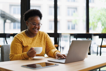 Smiling african american freelancer is working on a laptop and drinking coffee while sitting at a table in a cafe, she is wearing eyeglasses and a yellow sweater