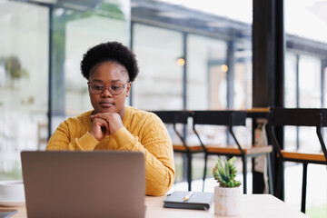 Young black businesswoman with eyeglasses concentrating on her laptop, working remotely in a bright and stylish cafe, demonstrating modern work practices and the gig economy