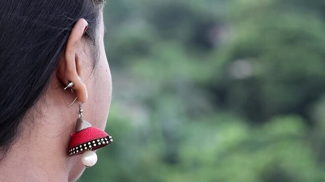 Elegant close-up of a Bangladeshi woman's ear with intricate red jhumka earrings crafted from silk thread. The timeless jewelry is set against a serene, blurred nature background.