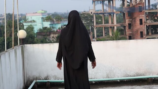 A Bengali woman in a black burqa and hijab stands on a rooftop, gazing down from the edge. Back view, capturing a sense of solitude and contemplation.