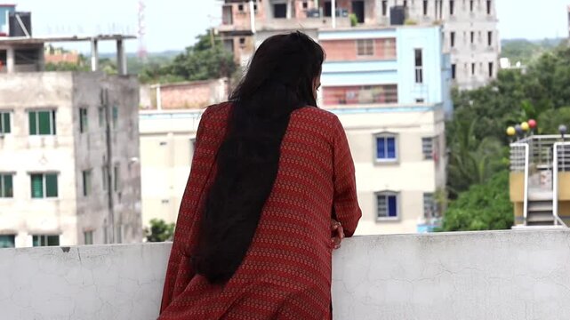A Bengali woman in a beautiful salwar kameez, standing alone on a rooftop. Her long, flowing black hair moves in the wind as she gazes at the scenery, a peaceful moment captured from the back.