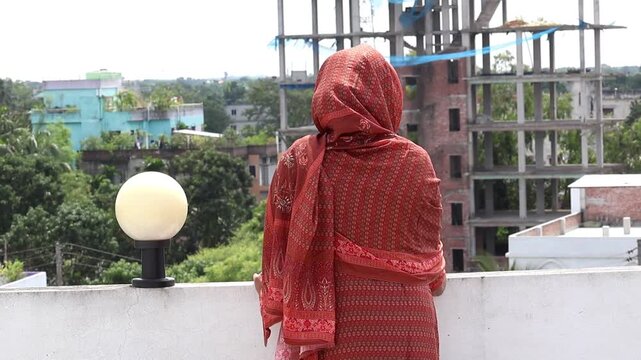 A Bengali Muslim woman in a salwar kameez and veil stands alone on a sunlit rooftop, enjoying the fresh morning air. The peaceful moment is captured from a back view.