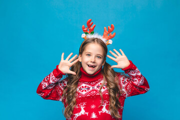 Cute little girl wearing a red Christmas sweater decorated with reindeer and a headband with reindeer antlers smiles and poses against a blue background. Santa Claus helper concept. High quality photo