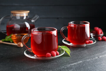 Aromatic raspberry tea in glass cups, teapot, berries, green leaves and honey on black table, closeup