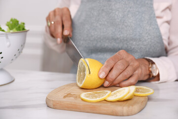 Senior woman cutting lemon at white marble table indoors, closeup