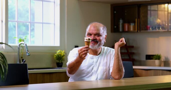 Senior Indian man eating ice cream in cone with delight at home, Asian elderly foodie enjoying cold dessert with childlike joy in modern kitchen, happily savoring frozen creamy treat in summer