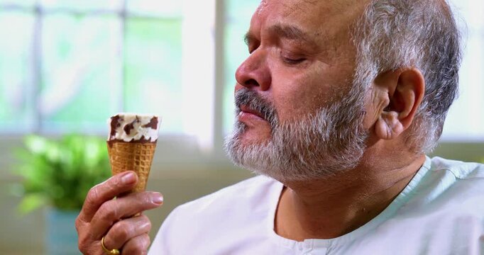 Senior Indian man eating ice cream in cone with delight at home, Asian elderly foodie enjoying cold dessert with childlike joy in modern kitchen, happily savoring frozen creamy treat in summer