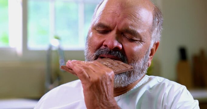 Senior Indian man secretly eating chocolate brick in modern kitchen, Asian elderly foodie sneaking thick chocolate slab with mischievous expression, enjoying sweet treat alone at home