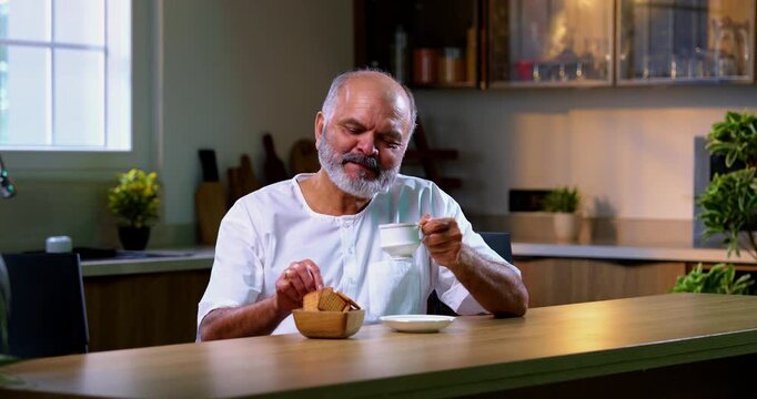 Senior Indian man eating tea biscuit in modern kitchen, Asian elderly man enjoying morning snack quietly, dipping crunchy biscuit into hot chai and savoring peaceful breakfast moment at home