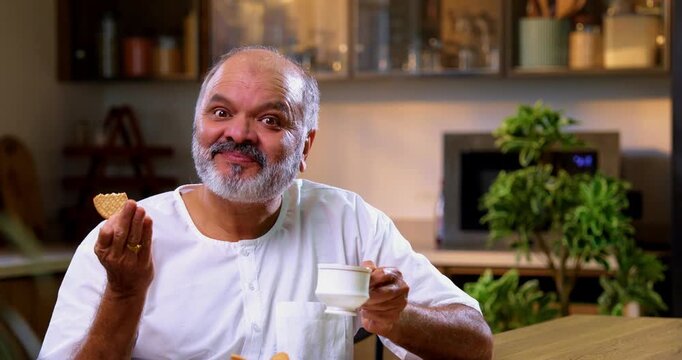 Senior Indian man eating tea biscuit in modern kitchen, Asian elderly man enjoying morning snack quietly, dipping crunchy biscuit into hot chai and savoring peaceful breakfast moment at home