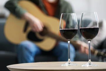 Glasses with red wine on table and man playing guitar indoors, selective focus