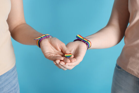 LGBT concept. Women in rainbow wristbands with heart shaped pin holding hands on light blue background, closeup