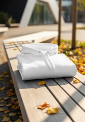 A clean, folded white hoodie sits on a sunlit wooden bench surrounded by fallen autumn leaves, offering a cozy and stylish apparel mockup scene.