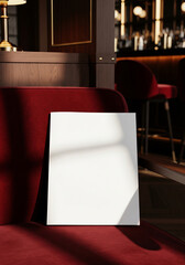 A blank white square canvas or sign mockup rests against a luxurious red velvet couch in a dimly lit, upscale bar or lounge setting.