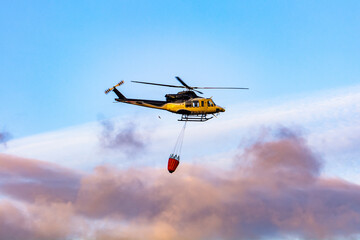 Yellow firefighting helicopter flying with water bucket during wildfire operation against colorful cloudy sky at sunset