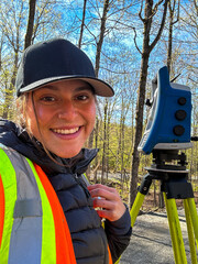 A young beautiful woman wearing a high visibility vest smiles at the camera with a transit for surveying and a forest in the background.