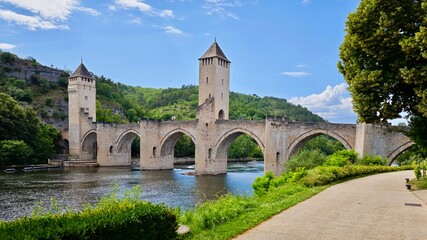Panoramic view of famous medieval Valentre bridge in Cahors, France. Medieval stone bridge with three towers spans a calm river, surrounded by lush greenery and rolling hills. France wine route. 
