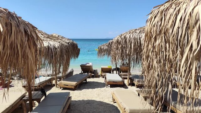 Video of straw-thatched umbrellas and wooden loungers at a beach bar in Alimos on Attica riviera, offering a serene seaside escape under the bright blue sky.