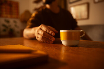 Close-up of a hand reaching for a coffee cup on a wooden table in a cozy, softly lit room.