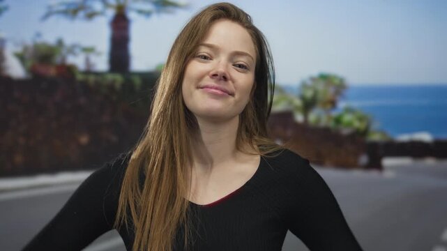 Woman points finger on seaside promenade with palm trees and ocean in view and wears a warm smile; friendly greeting.