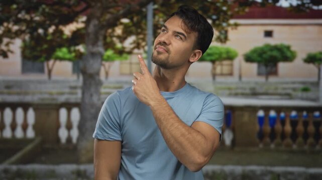 Young hispanic man outdoors pointing with different expressions in a serene garden setting, blending curiosity and contemplation amidst lush green surroundings and historic architecture.
