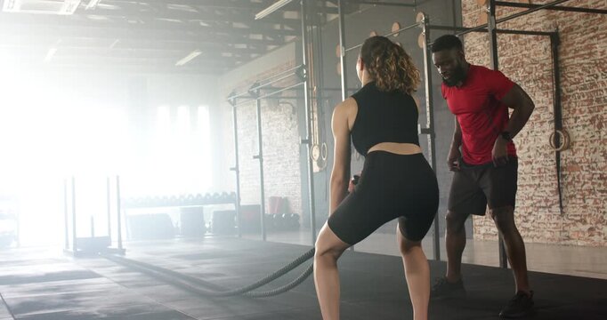 African American trainer guiding White woman doing battle rope waves for strength at gym