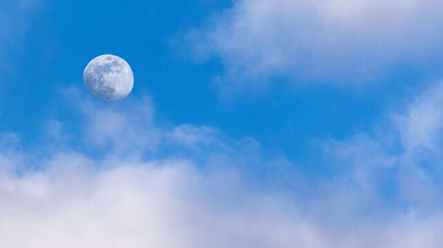full moon against sunny blue sky surrounded by clouds