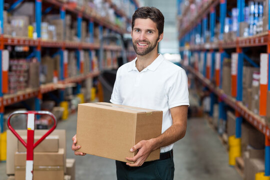 Warehouse worker is holding cardboard box in central aisle between steel racks and red pallet jack