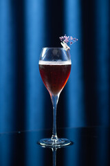 A wine glass filled with a dark red cocktail, decorated with a small purple flower, placed on a reflective black surface with a dark blue background