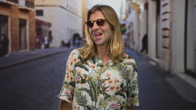Man with long blonde hair wearing sunglasses and a colorful floral shirt smiling on a sunlit street; vacation bliss.