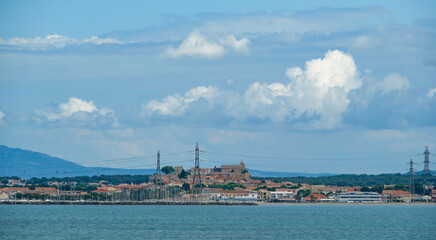 Coastal town with a hilltop historic church, marina with sailboats, and power lines under a blue sky with dramatic clouds