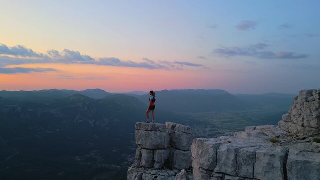 Determined runner triumphantly stands atop a rugged cliff, overlooking a vast mountainous landscape at sunset. Captures endurance, achievement, and joy of conquering nature's challenges, wide shot.