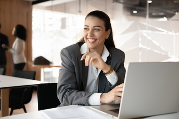 Fototapeta premium Creative thinking and visualization. Cheerful young businesswoman sit by laptop at workplace desk look away with dreamy smile create new solution reflect on future goals take mindful break. Copy space