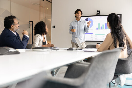 Business development briefing. Young man ceo boss stand by table in conference room lead strategy planning session for multicultural staff use visual data on tv screen to guide team support discussion