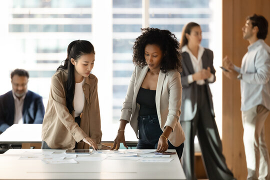 Productive paperwork. Focused multicultural team two young female project managers colleagues develop creative startup review financial report study statistical data in paper documents on office desk - Powered by Adobe
