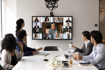 Multinational corporation hybrid meeting. Group of businesspeople top executives from different ethnic backgrounds gather around conference table talk to remote teammates via videocall on big screen