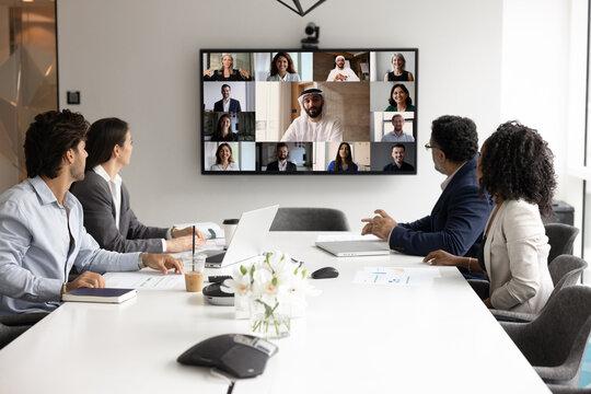 International business collaboration. Multiethnic businesspeople corporate employees gather at boardroom for hybrid virtual conference with colleagues join group meeting via videocall on large screen