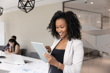 Hybrid teamwork. Cheerful young African businesswoman communicate on digital tablet stay connected online handle electronic paperwork while coworkers engaged in strategy discussion at conference desk