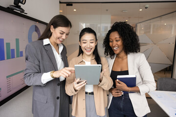 Professional friendship and successful work. Multiethnic group of 3 young businesswomen having fun together look at tablet pc explore new ideas in casual office setting discuss job with joy laughter