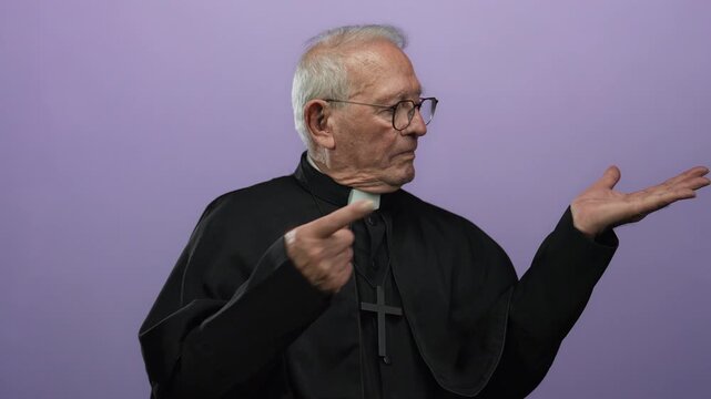 Senior priest in black robe gestures with open hand against isolated purple background.